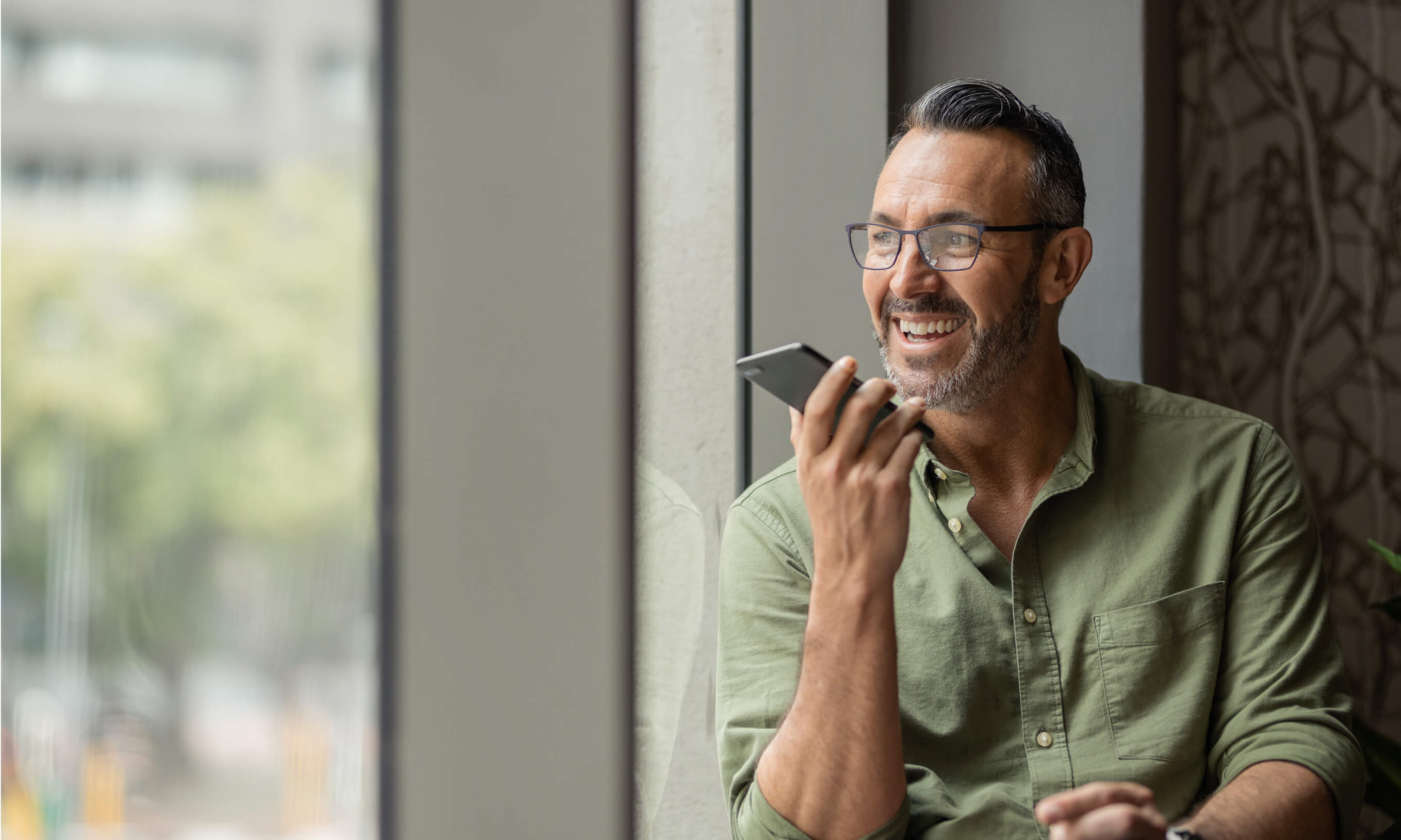 A man smiling holding a phone against a funky dark wallpaper and a large window.